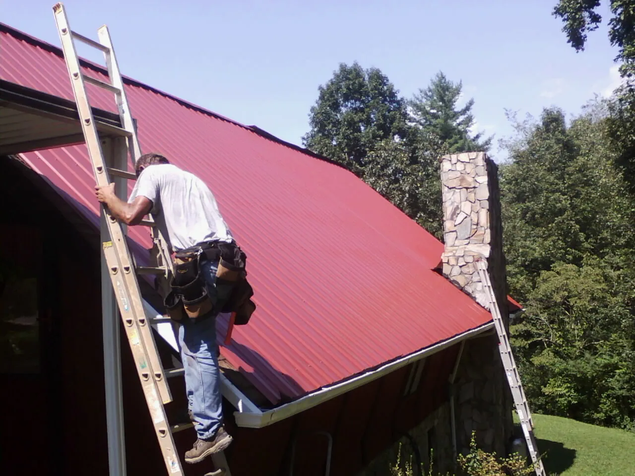Roofer on a ladder beside a red metal roof and stone chimney — Soffit & Fascia Repair by Roofing Brothers in Livermore Falls