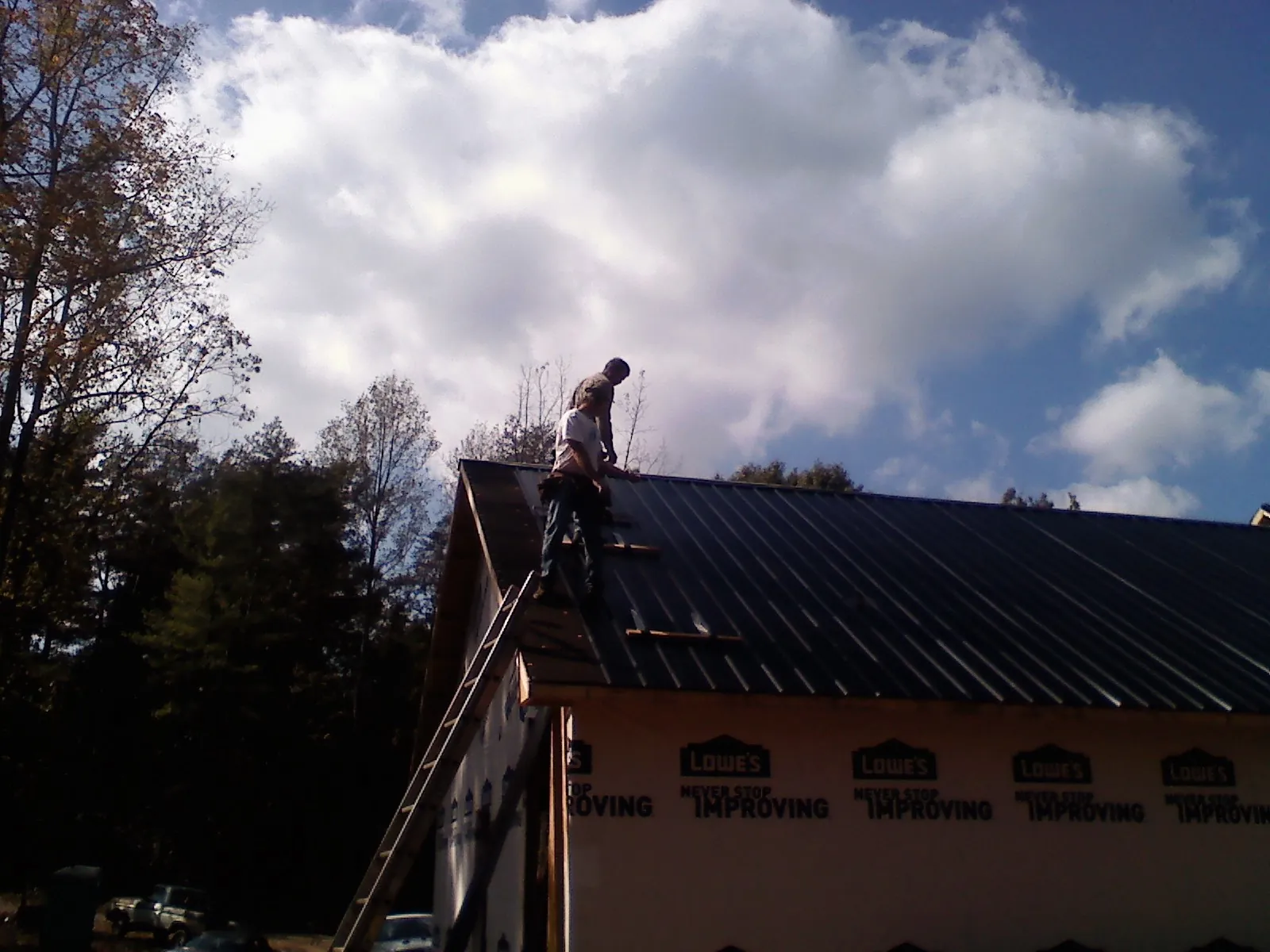 Roofer fastening dark metal panels high against a cloudy sky, from a Roof Inspection job in Livermore Falls